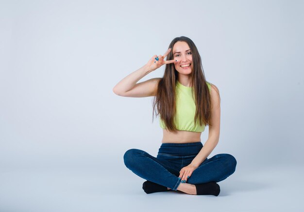 Jeune fille souriante montre deux gestes en s'asseyant sur le sol sur fond blanc