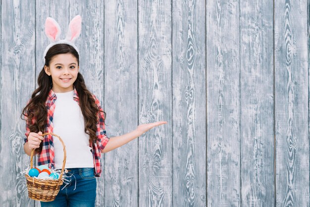 Jeune fille souriante avec des oreilles de lapin tenant un panier d&#39;oeufs de Pâques présentant contre un bureau en bois gris