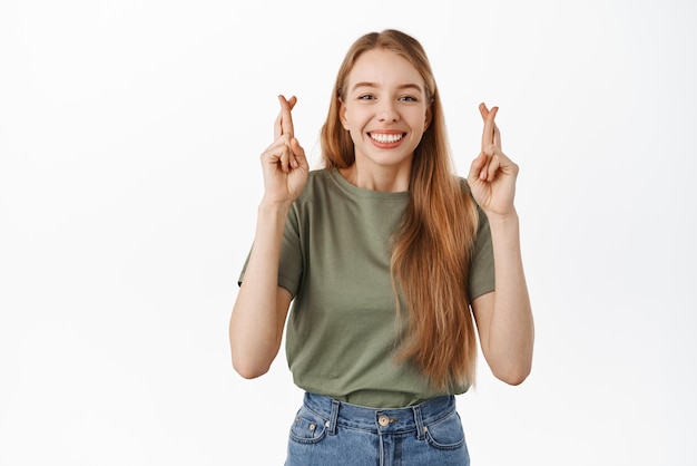 Une jeune fille souriante pleine d'espoir croise les doigts pour la bonne chance et sourit heureuse en attendant de bonnes nouvelles en priant anticipant la victoire debout sur fond blanc