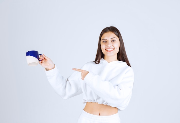 jeune fille souriante pointant sur une tasse sur fond blanc-gris.