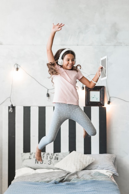 Jeune fille souriante portant un casque sautant par-dessus le lit avec tablette numérique