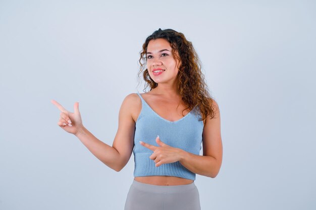 Jeune fille souriante regarde ailleurs en pointant vers la gauche avec les index sur fond blanc