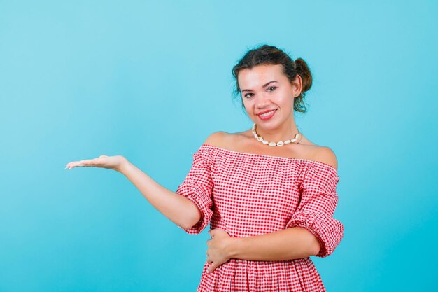 Jeune fille souriante regarde la caméra en ouvrant la main à droite sur fond bleu