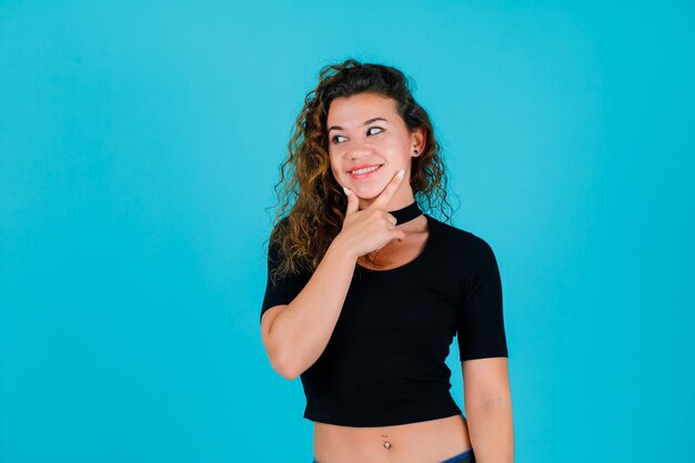 Jeune fille souriante regarde à gauche en mettant les mains sur le menton sur fond bleu