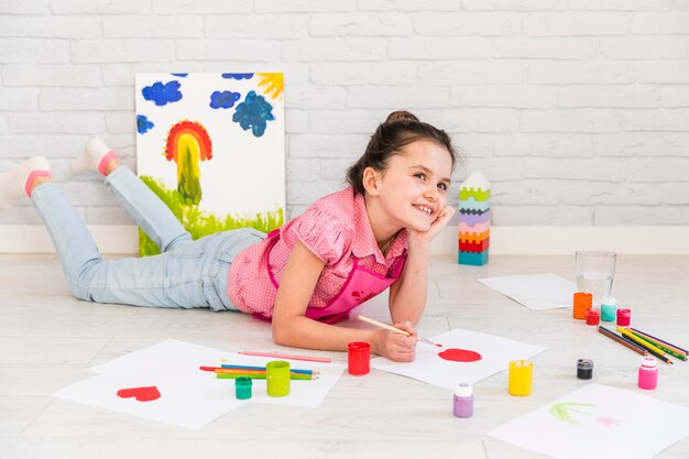 Jeune fille souriante se trouvant sur la peinture de sol sur papier blanc avec un pinceau rouge