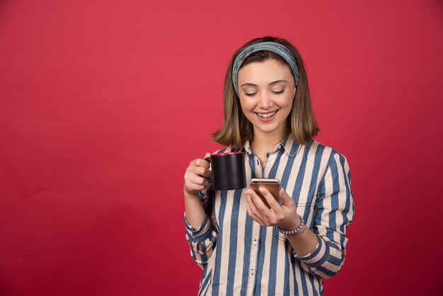 Jeune fille souriante avec une tasse de thé vérifiant le téléphone portable