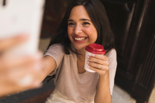 Jeune fille souriante tenant une tasse de café à emporter prenant selfie depuis un smartphone