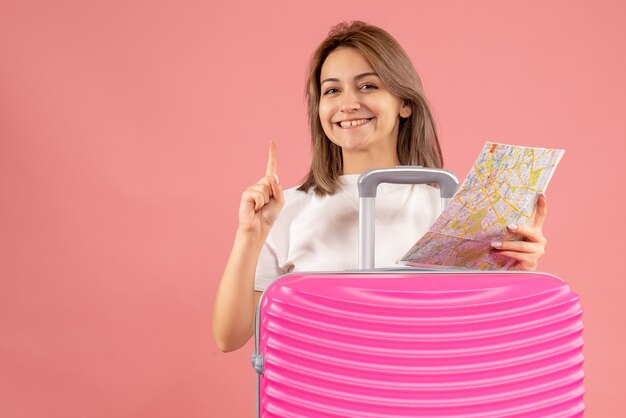 jeune fille souriante avec une valise rose tenant une carte pointant le doigt vers le haut