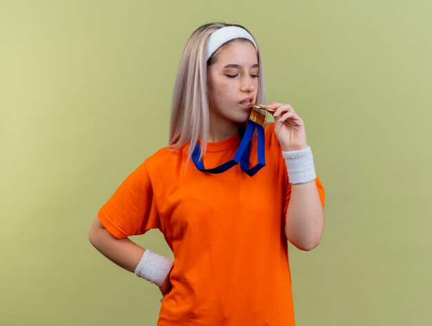 Une jeune fille sportive caucasienne confiante avec des bretelles portant un bandeau et des bracelets fait semblant de mordre la médaille d'or