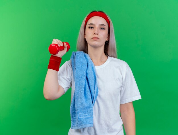 Jeune fille sportive caucasienne confiante avec des bretelles portant un bandeau et des bracelets avec une serviette sur l'épaule tient un haltère