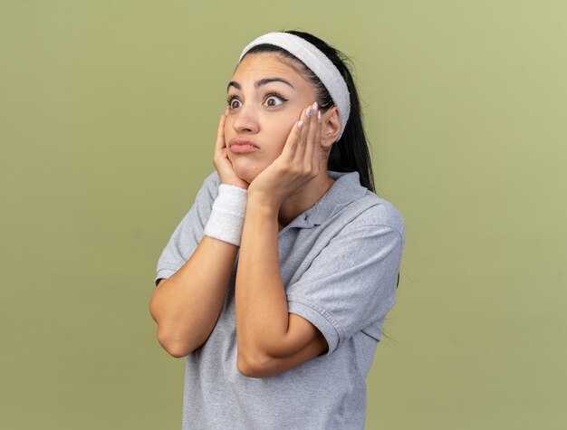 Jeune fille sportive caucasienne ignorante portant un bandeau et des bracelets debout en vue de profil en gardant les mains sur le visage en regardant de côté isolé sur un mur vert olive avec espace de copie