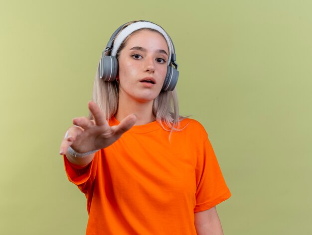 Jeune fille sportive caucasienne impressionnée avec des bretelles sur des écouteurs portant un bandeau et des bracelets qui s'étendent la main