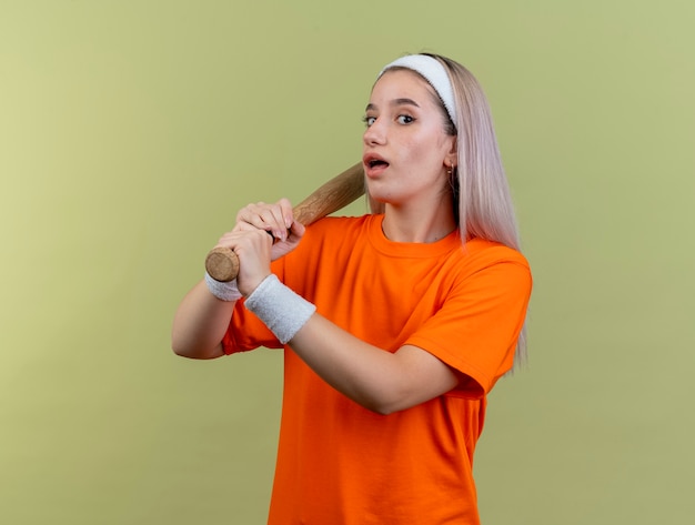 Jeune fille sportive caucasienne surprise avec des bretelles portant un bandeau et des bracelets tenant une batte de baseball