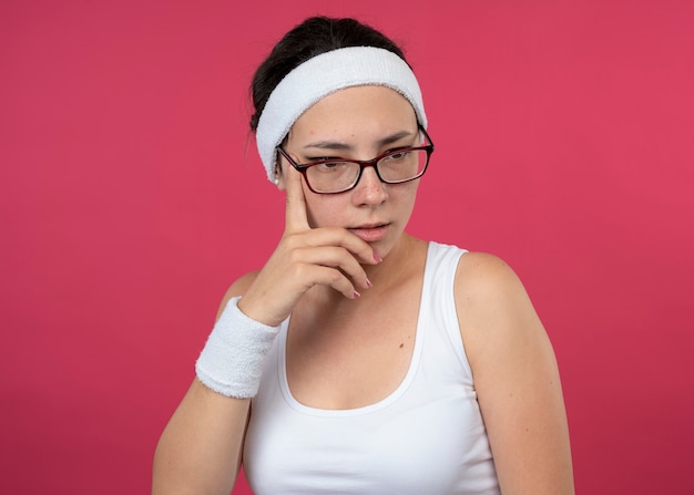 Une jeune fille sportive réfléchie dans des lunettes optiques portant un bandeau et des bracelets met la main sur le menton et regarde vers le bas