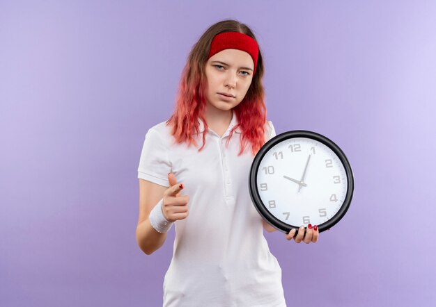 Jeune fille sportive tenant horloge murale avec visage sérieux pointant avec le doigt debout sur le mur violet