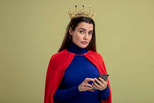 Jeune fille stricte de super-héros portant la couronne tenant le téléphone isolé sur vert olive