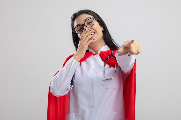 Jeune fille de super-héros caucasien portant des lunettes et un stéthoscope en riant en gardant la main sur la bouche à la recherche et en pointant la caméra isolée sur fond blanc avec espace de copie