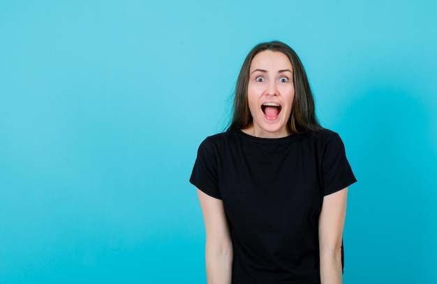 Une jeune fille surprise regarde la caméra en ouvrant grand la bouche sur fond bleu