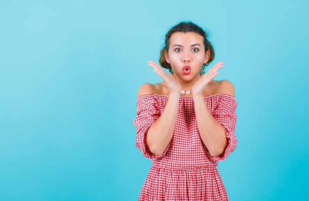 Une jeune fille surprise regarde la caméra en se tenant la main près des joues sur fond bleu