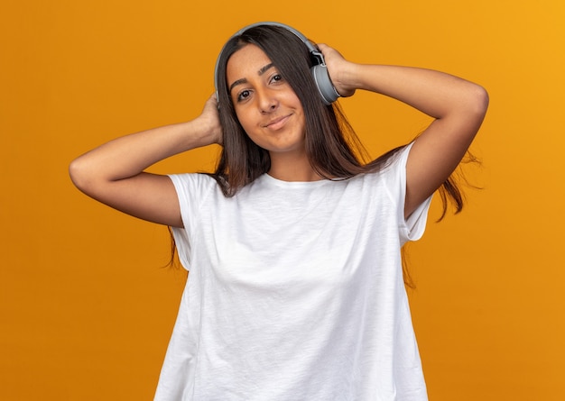 Jeune fille en t-shirt blanc avec un casque heureux et positif regardant la caméra souriante appréciant sa musique préférée debout sur fond orange