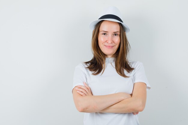 Jeune fille en t-shirt blanc, chapeau debout avec les bras croisés et regardant confiant, vue de face.