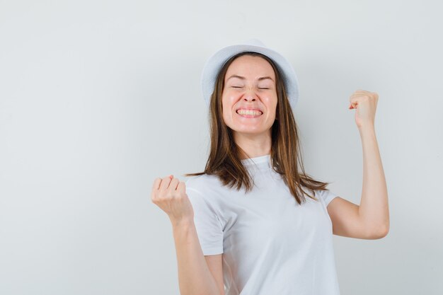 Jeune fille en t-shirt blanc, chapeau montrant le geste du gagnant et regardant heureux, vue de face.