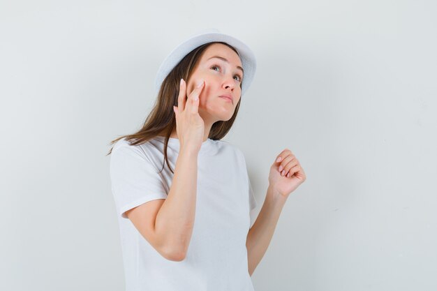 Jeune fille en t-shirt blanc, chapeau touchant la peau de son visage sur la joue, vue de face.