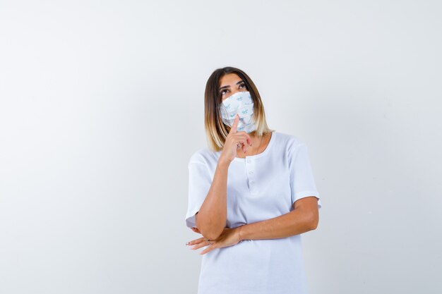 Jeune fille en t-shirt blanc, masque mettant l'index sous le menton, regardant vers le haut, pensant à quelque chose et regardant pensif, vue de face.