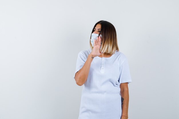 Jeune fille en t-shirt blanc, masque montrant un panneau d'arrêt, regardant ailleurs et regardant sérieusement, vue de face.