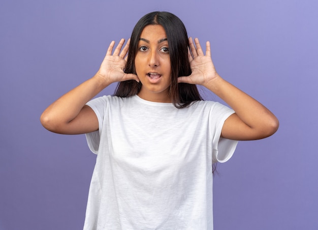 Jeune fille en t-shirt blanc regardant la caméra heureuse et surprise tenant les paumes ouvertes sur ses oreilles, debout sur fond bleu