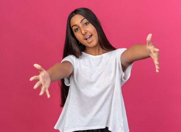 Jeune fille en t-shirt blanc regardant la caméra souriant joyeusement faisant un geste de bienvenue large ouvrant les mains debout sur fond rose