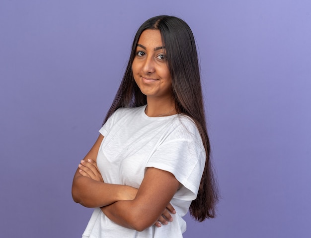 Jeune fille en t-shirt blanc regardant la caméra avec le sourire sur le visage avec les bras croisés debout sur bleu