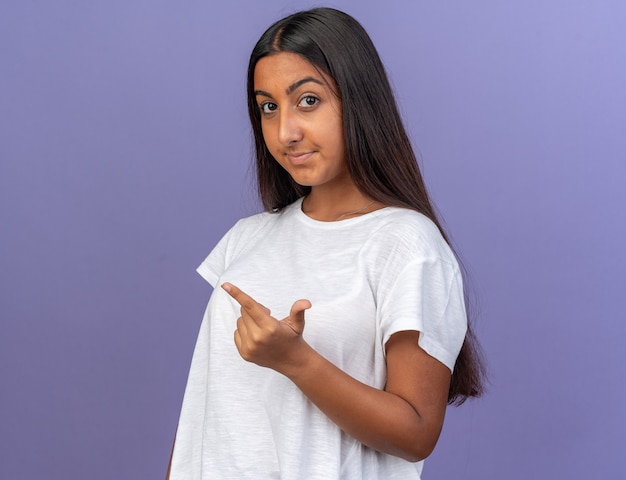 Jeune fille en t-shirt blanc regardant la caméra avec un sourire sur le visage montrant l'index faisant des gestes comme demander debout sur bleu