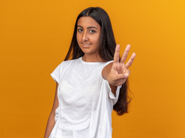 Jeune fille en t-shirt blanc regardant la caméra avec le sourire sur le visage montrant le numéro trois avec les doigts