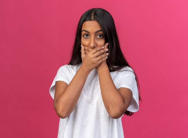 Jeune fille en t-shirt blanc regardant la caméra en train d'être choquée couvrant la bouche avec les mains debout sur fond rose