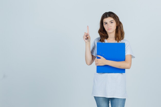 Jeune fille en t-shirt blanc tenant le dossier tout en pointant vers le haut et à la vue sensible, de face.