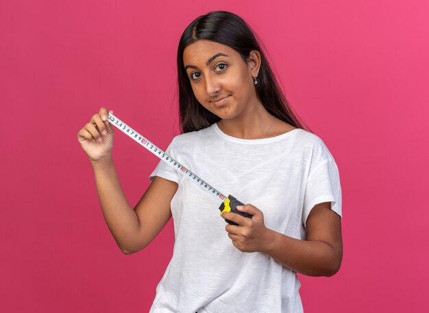 Jeune fille en t-shirt blanc tenant un ruban à mesurer regardant la caméra avec un sourire sur le visage heureux et positif