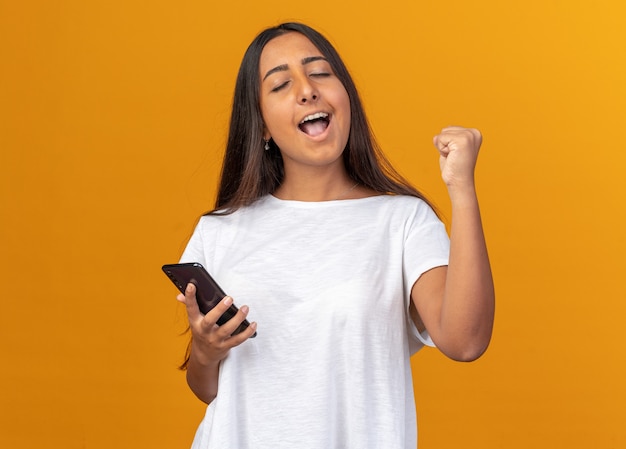 Jeune fille en t-shirt blanc tenant un smartphone heureux et excité, serrant le poing debout sur fond orange