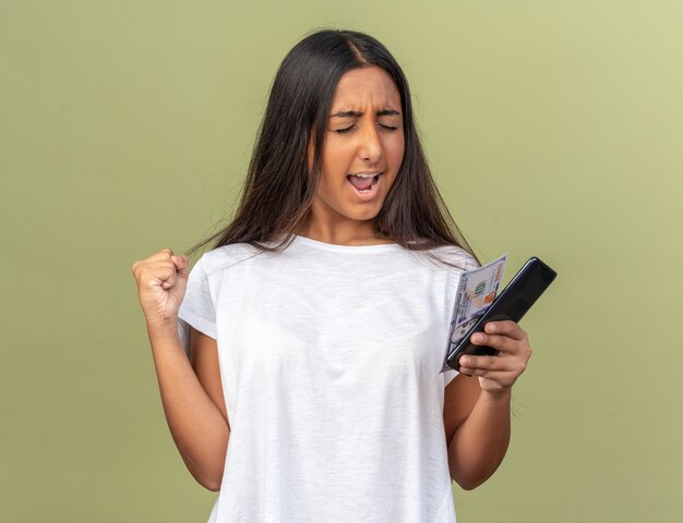 Jeune fille en t-shirt blanc tenant un smartphone heureux et excité serrant le poing debout sur le vert