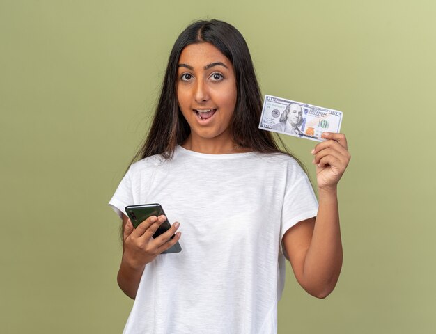 Jeune fille en t-shirt blanc tenant un smartphone montrant de l'argent en regardant la caméra heureuse et surprise