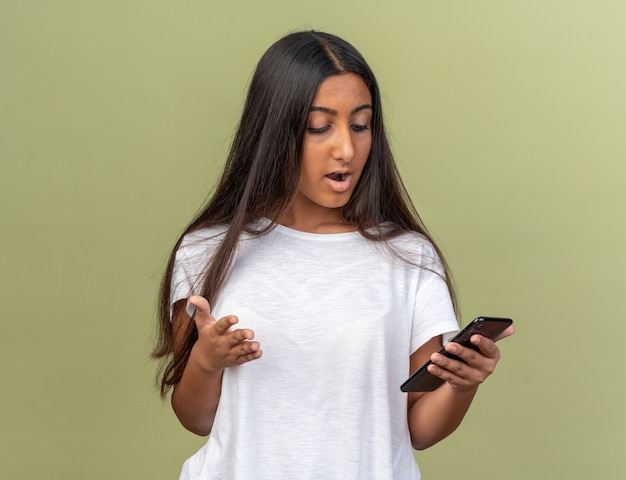 Jeune fille en t-shirt blanc tenant un smartphone le regardant confus et très anxieux debout sur green