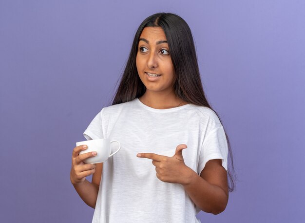 Jeune fille en t-shirt blanc tenant une tasse blanche pointant avec l'index en regardant de côté avec le sourire sur le visage