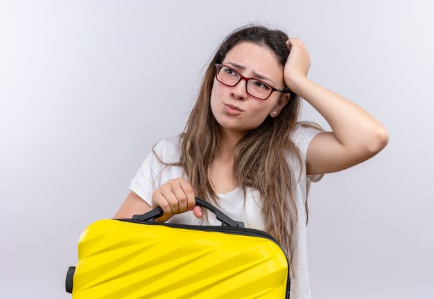 Jeune fille en t-shirt blanc tenant une valise de voyage à côté confus avec la main sur la tête