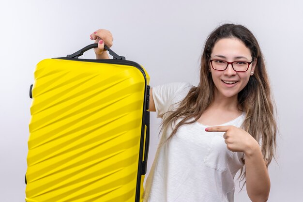 Jeune fille en t-shirt blanc tenant une valise de voyage pointant avec le doigt dessus en souriant joyeusement