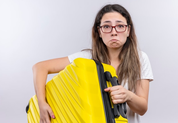 Jeune fille en t-shirt blanc tenant une valise de voyage regardant la caméra avec une expression triste avec un visage malheureux