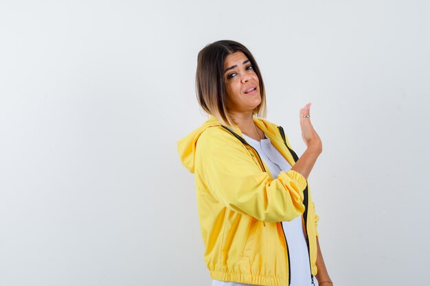 Jeune fille en t-shirt blanc, veste jaune levant la main comme agitant, saluant quelqu'un et regardant aimable, vue de face.