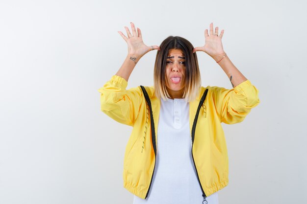 Jeune fille en t-shirt blanc, veste jaune tenant la main près de la tête, tirant la langue et à la vue amusée, de face.