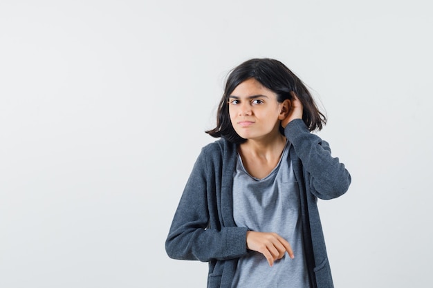 Jeune fille en t-shirt gris clair et sweat à capuche zippé gris foncé levant la main près de l'oreille comme essayant d'entendre quelque chose et à la recherche concentrée