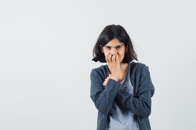 Jeune fille en t-shirt gris clair et sweat à capuche zippé gris foncé mettant la main sur la bouche, posant à la caméra et regardant mignon