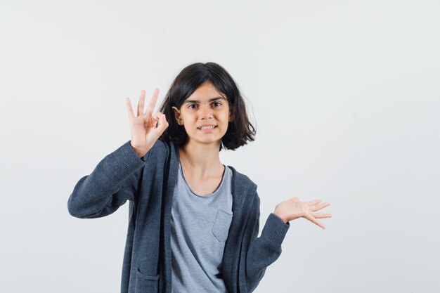 Jeune fille en t-shirt gris clair et sweat à capuche zippé gris foncé montrant le signe ok et étirant la main et à mignon